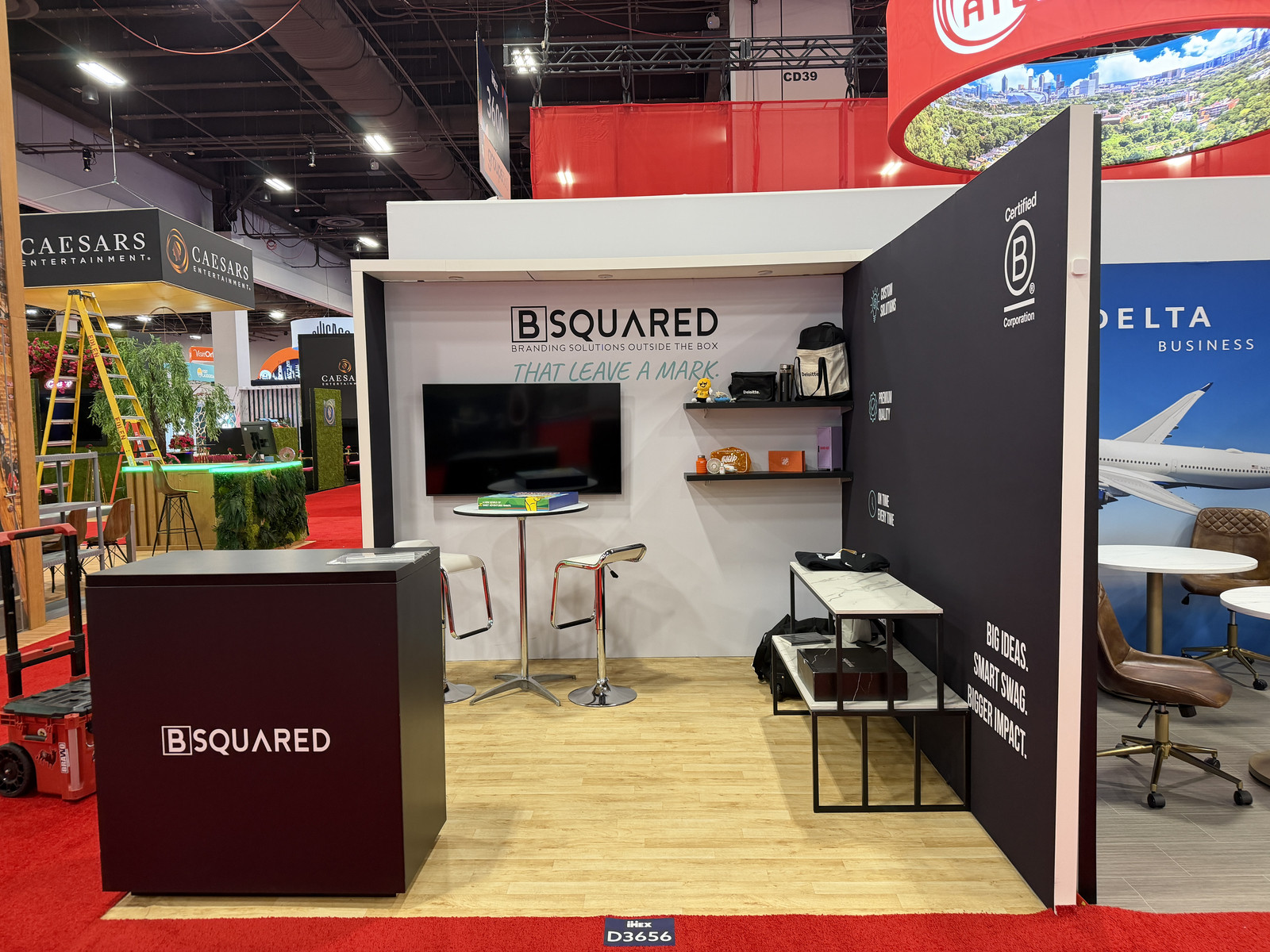 BSQUARED 10x10 trade show exhibit at IMEX 2026 featuring branded wall graphics, a reception counter, product display shelves, a mounted screen, and a small meeting area.