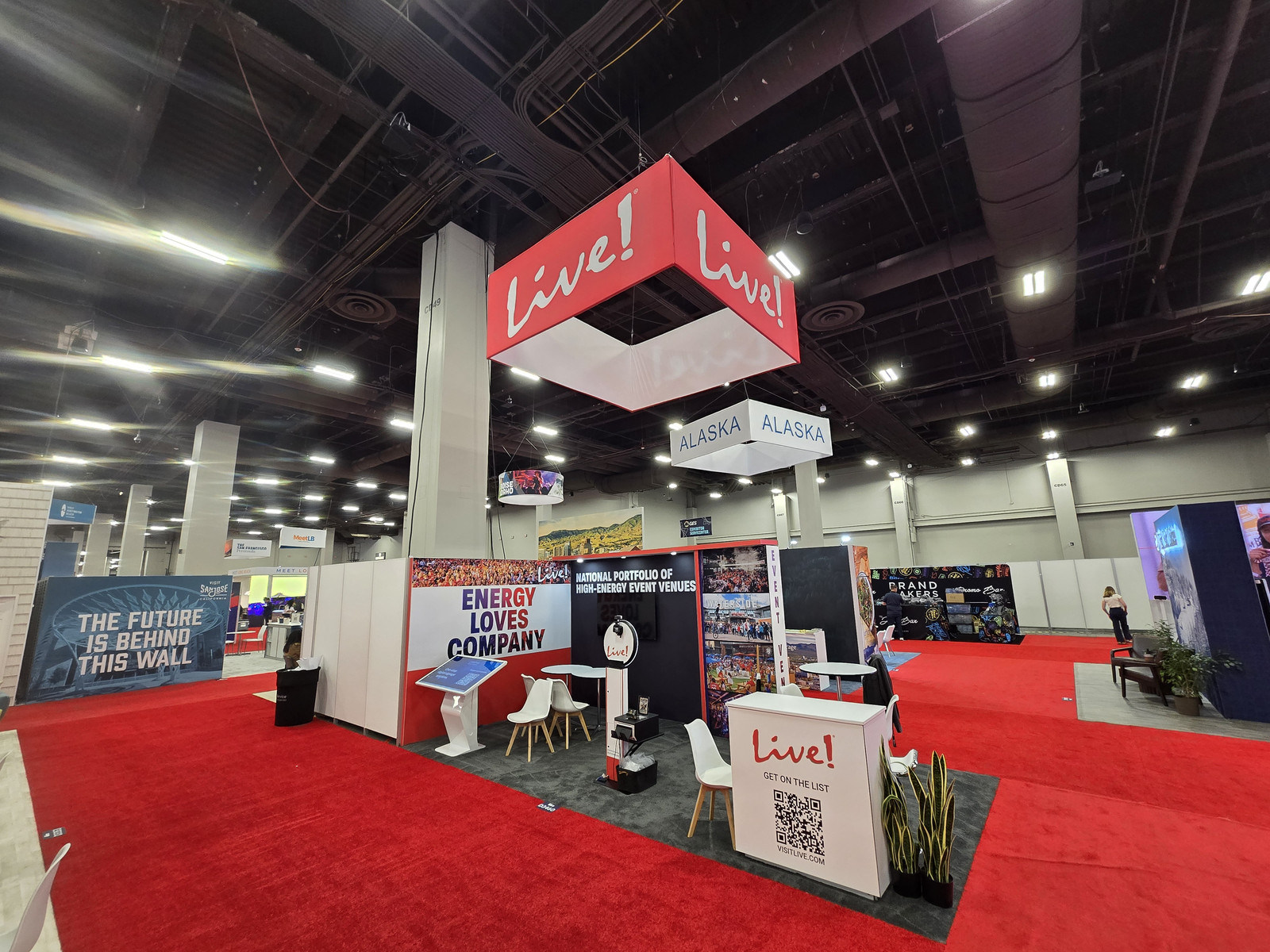 Live! Hospitality 10x20 trade show exhibit at IMEX 2026 featuring a suspended red Live! sign, event venue graphics, interactive displays, meeting tables, and a branded reception counter.