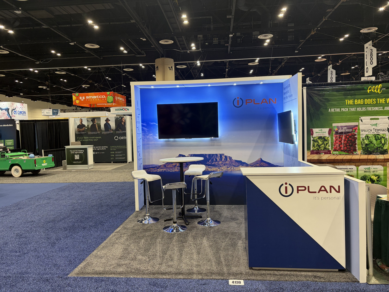 iPlan Global 10x10 trade show exhibit at the 2026 Global Produce & Floral Show featuring blue illuminated wall graphics, mounted screens, a branded reception counter, and meeting stools.